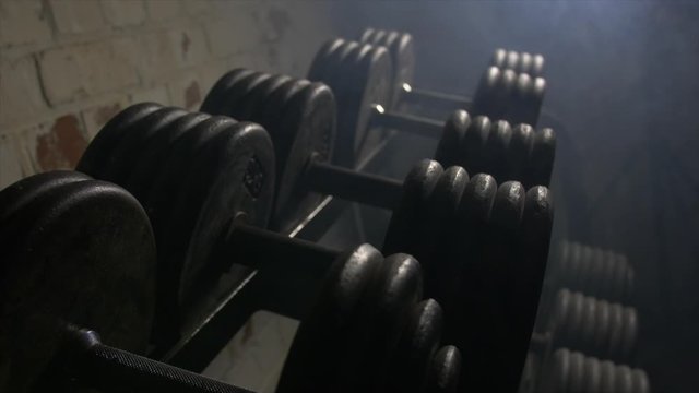 Close Up Pan Across Of Weights Lined Up On Weight Rack In Industrial Gym Old Style Old Fashioned Under Ground Gym. Paint Work Scratched Off Smoke Passing Over Dark Lit Room
