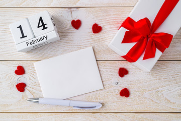 Empty greeting card, gift box with red ribbon bow and perpetual calendar with date 14 february on a white wood textured surface. Romance and love