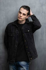 Obraz premium Studio fashion portrait of attractive young man in black jacket and blue jeans standing over grey textured wall. Guy correct hair with his hand