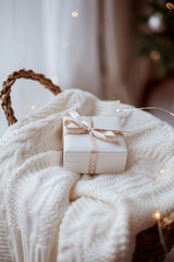 Fototapeta na wymiar small white gift box with ribbon on a knitted sweater, wicker basket, Christmas lights.