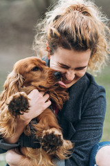 Cute cocker spaniel on hands of attractive happy young curly woman. The dog licks the owner