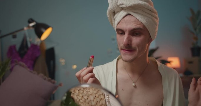 Close up of man travesty diva sitting, putting pink lipstick and getting ready for show. Young man transvestite and looking in mirror while putting on make up and moving head.