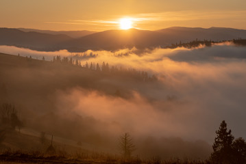 Carpathians, dawn in the valley, the fog is illuminated by warm sunlight, in the foreground trees, haystacks, in the background mountains and hills.