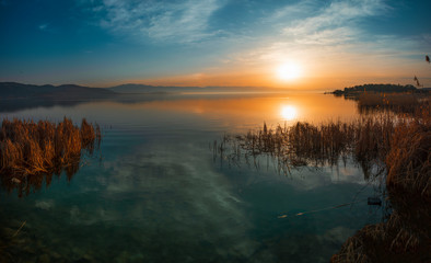 Beautiful sunrise on the lake and mountains. Bright sun rays with fog reflected on the lake with the reeds on the shore.