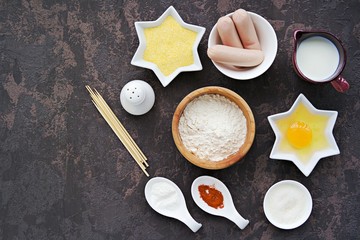 Ingredients for making corn dogs on a dark concrete background : sausages, corn flour, wheat flour, egg, sugar, baking powder, salt, wooden skewers, paprika, milk.