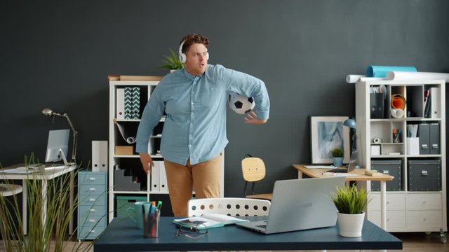 Playful guy wearing headphones is having fun with football in office enjoying music during work break. Entertainment, modern devices and people concept.