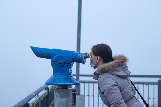Young woman standing at city viewpoint, wearing mask to protect her from smog, looking through telescope in thick fog. Conceptual photo about ecology and air pollution.