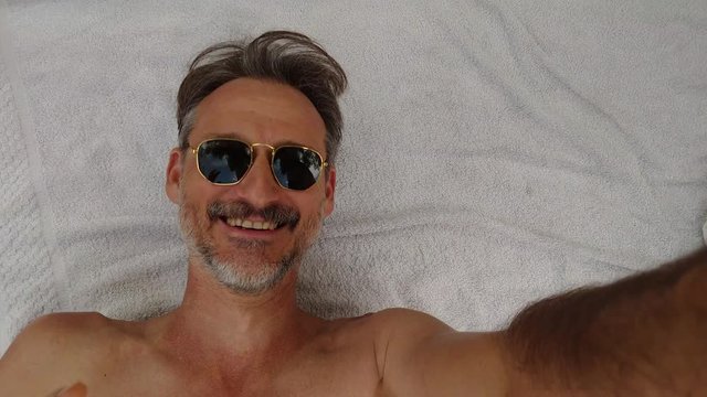 Close-up portrait of the happy man with glasses, waving at the smarphone's selfie camera on the beach. Background towel