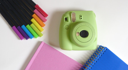 Green Instant Camera with Multicolored Brush Pens on a White Background with a Blue Notebook and Pink Binder