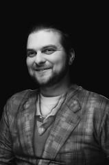 black and white low-key portrait of cheerful handsome bearded stylish elegant man in plaid blazer smiling and looking away on dark background