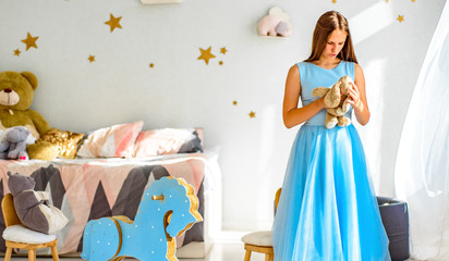 portrait of young teenager brunette girl with long hair in blue dress in her bedroom and play with children’s toy