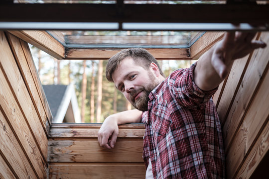 Handsome Bearded Man Stands Near An Open Skylight In The Attic.