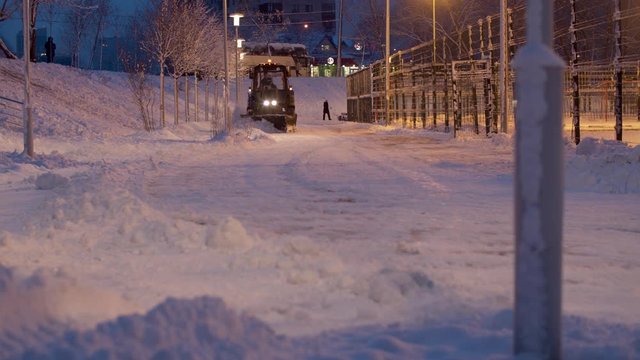Tractor grader removing snow from the road in the snow covered park. Winter evening. Light of lanterns, luminous windows in the houses. Road service in work