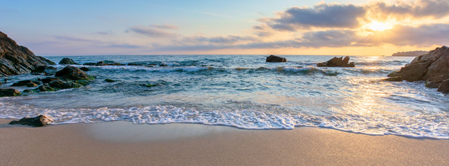 Fototapety  wschód słońca na plaży. piękna letnia sceneria. skały na piasku. spokojne fale na wodzie. chmury na niebie. szeroki widok panoramiczny