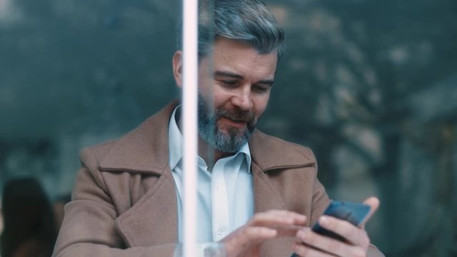 Close up rotation view of gorgeous middle aged beared business man with beige coat uses phone drinks coffee at cafe texing message working successful people relaxing new technologies communicating