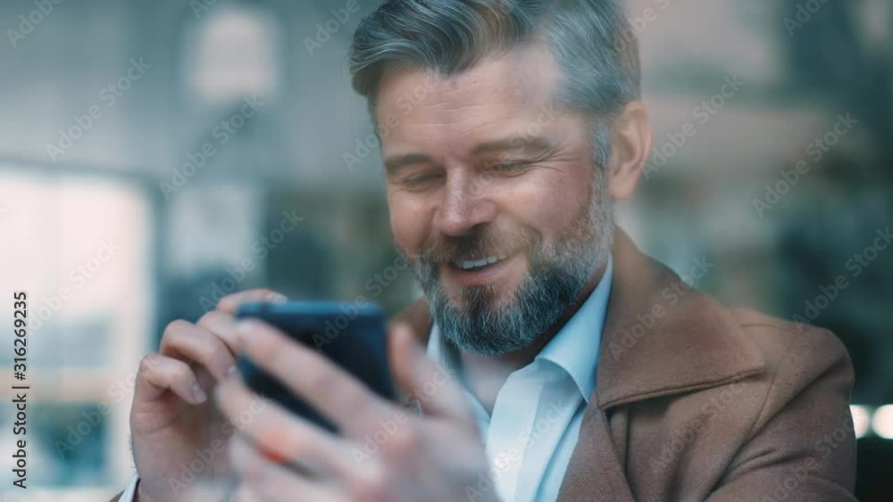 Wall mural portrait of gorgeous smiling middle aged beared business man uses phone drinks coffee at cafe texing