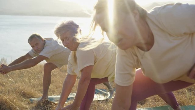 Group of senior people doing yoga exercises at nature. Close-up portrait elderly woman outstretched hands folded in gesture while standing in row with friends in sunset shining at picturesque view