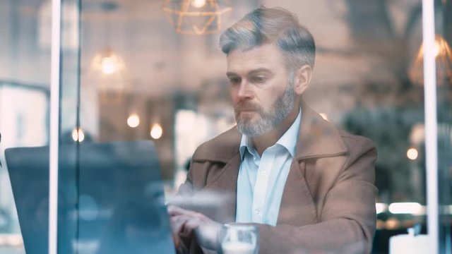 Close up view through the window of caucasian gorgeous middle aged man with grey hear works in coffee shop Man uses laptop business and entrepreneurship successful confident social network apps laptop