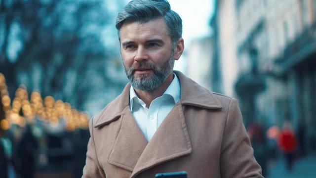 Attractive mature european man with grey hair walks down the street drinks coffee uses phone texting message feels happy smile city lights on background slow motion close up portrait business