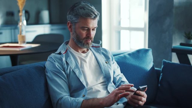 Close up view of cheerful middle aged man with grey hair sitting on the sofa using phone smile in the modern apartment texting message scrolling tapping technology isolated lifestyle slow motion