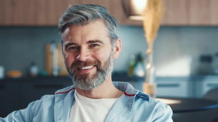Portrait of happy middle aged man sitting on sofa looking to the camera and smiling in modern room close up slow motion - Powered by Adobe