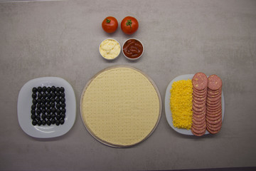 pizza preparation process top view: two round blanks of pizza dough with salami olives, cheese on plates with two tomatoes and two sauces next to it on a gray table with texture
