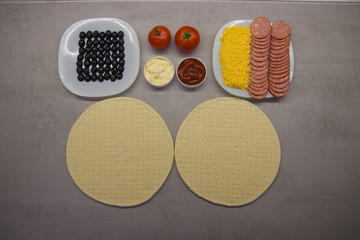 pizza preparation process top view: two round blanks of pizza dough with salami olives, cheese on plates with two tomatoes and two sauces next to it on a gray table with texture