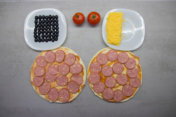 two round pizzas with salami cheese and olives tomatoes and sauce and on a gray table with texture top view