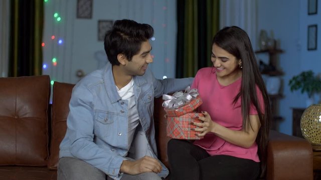 Handsome young guy gifting a Birthday or Valentines Day present to his girlfriend. Pretty Indian girl hugging after getting a colorful gift box from his romantic boyfriend while celebrating Valenti...