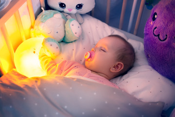 Baby sleeps in bed with lighting moon and soft toys in the night