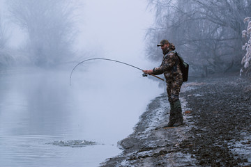 Fisher man fishing with spinning rod on a river bank at misty foggy winter, spin fishing, prey fishing