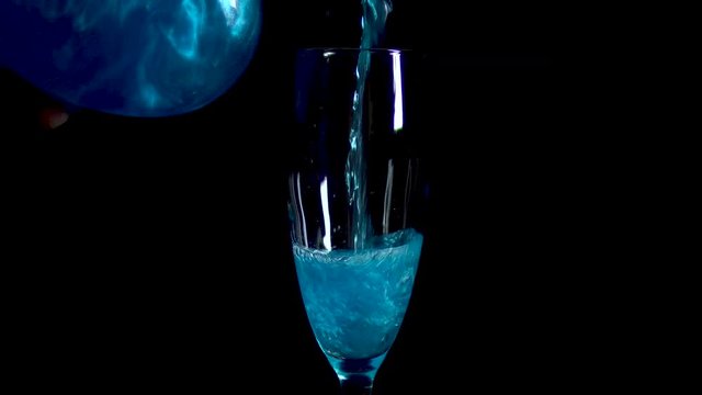 A blue drink is poured into a glass on a black background. The elixir spins and shimmers. Potion of love is isolated on a black background.