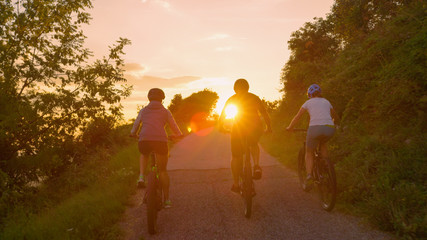 LENS FLARE: Unrecognizable cyclists pedal along the empty road at golden sunset.