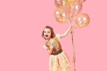 Happy little girl in glittering dress looking at camera on pink background and holding balloons.