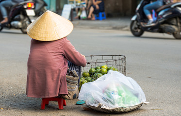 A local Vietnamese woman in a traditional Vietnamese hat sits on the street and sells fruits. Next to her, motorbikes pass along the street.