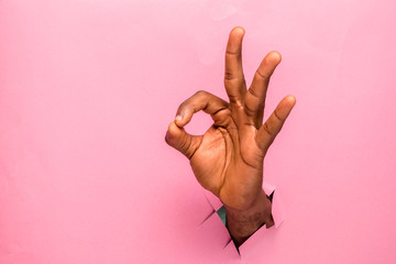 black person hand through a hole in a cardboard making ok sign on pink background