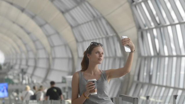 woman taking selfie picture with mobile phone at airport terminal in travel. portrait of young female in international hall holding coffee cup. concept airplane, photo, waiting room