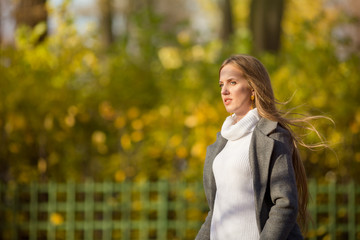 Beautiful girl with light brown hair walks in the autumn park. Attractive young woman in a gray coat and white sweater looks away. Fall season. Amazing autumn mood.