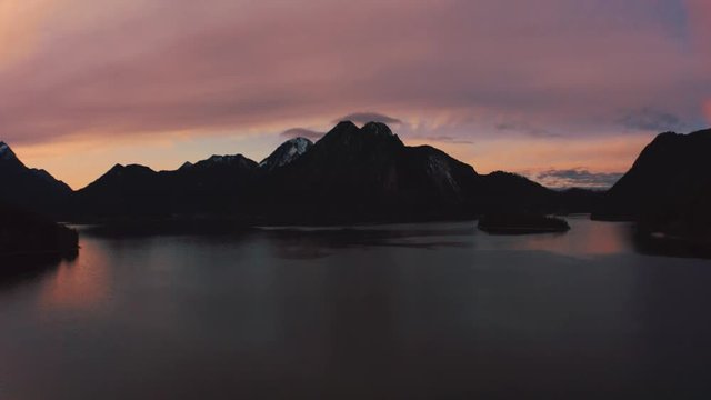 Anamorphic 4K Aerial Drone footage of the famous, scenic and idyllic Bavarian lake Walchensee in southern Germany in the alps. The sunset is coloring the clouds above the snowy mountain tops red.