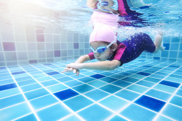 Underwater young little cute girl is swimming in the swimming pool with her swimming teacher. Seen under water while she is diving forward ahead of her. Swimming kid in the pool concept.