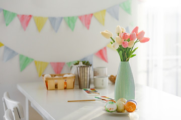 Easter home decorations, multi-colored flags, tulips and eggs on a white table on a bright clear day in a snow-white kitchen