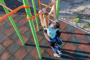 Young fit athlete working out at outdoor gym making dipping pull ups exercise using dip rings with his girlfriend hanging on his lap.