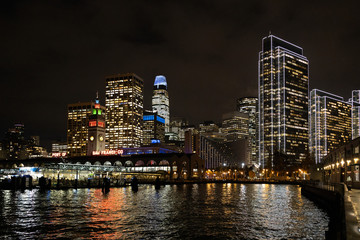 Fototapeta na wymiar San Francisco port illuminated at night, against the backdrop of impressive skyscrapers with light and reflection in the water, view from the side of the San Francisco Bay.