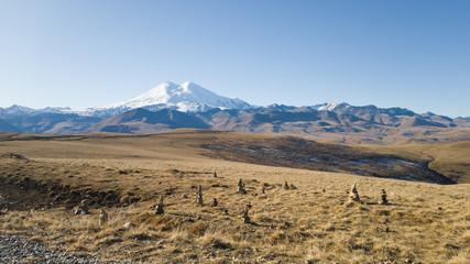 Fototapeta na wymiar Autumn landscape. View of the dry yellow field and the beautiful mount Elbrus in the Caucasus. Pyramids of stones in the foreground. Travel to Russia