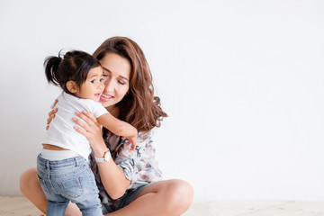 Young happy mother holds her charming mixed-race daughter and poses on a white wall background. Concept of a loving and caring family. Advertising space