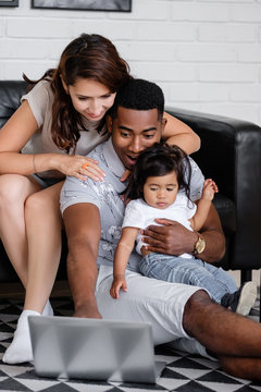 Charming mixed race family african-american father asian mother and charming little daughter watching together fascinating family movie using laptop and high speed internet sitting on sofa at home