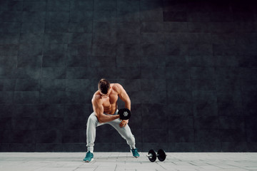 Handsome muscular caucasian blond shirtless man lifting dumbbell while standing in front of dark background.