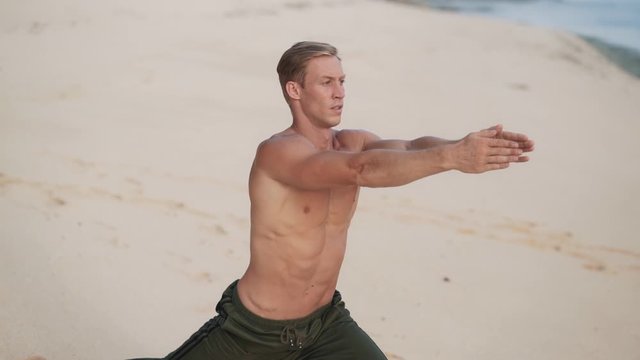 Man with muscular body doing yoga exercises on beach at sunset, slow motion.