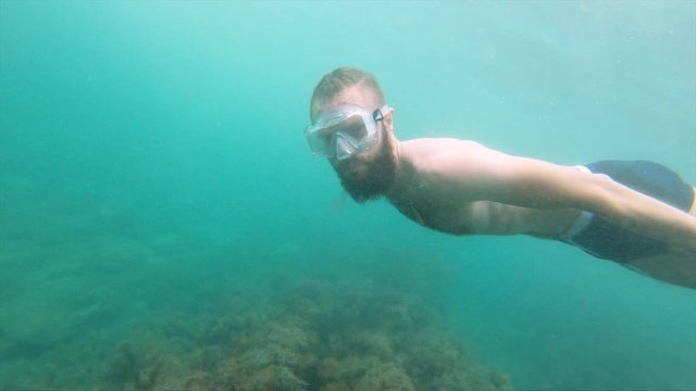 A thin man with a beard in a mask swims underwater, waving his arms. Close-up.