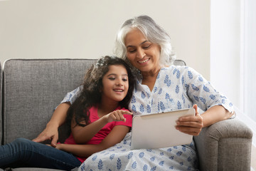 Portrait of a grand-daughter and grandmother sitting on the couch at home and looking at tablet. 	